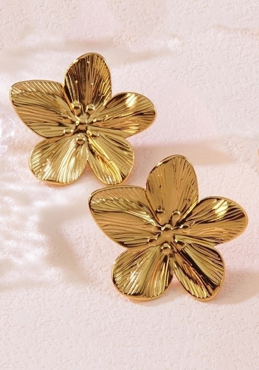 A pair of shiny, goldfarben Edelstahl Ohrstecker by Princess Goes Hollywood, shaped like five-petal flowers with textured details, rests on a soft pink surface. The lighting enhances their reflective finish and intricate design.