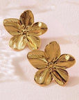 A pair of shiny, goldfarben Edelstahl Ohrstecker by Princess Goes Hollywood, shaped like five-petal flowers with textured details, rests on a soft pink surface. The lighting enhances their reflective finish and intricate design.