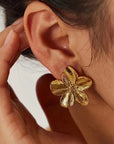 Close-up of a person’s ear adorned with gold floral stud earrings by Princess Goes Hollywood. Their hand gently touches the face, while dark hair is pulled back to highlight the shiny gold stainless steel earring.