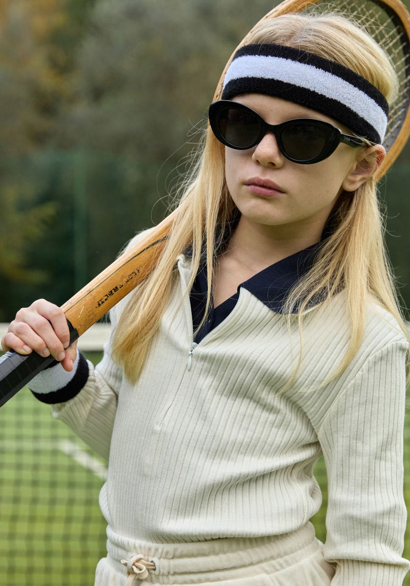 A girl with long blonde hair wears Okkia black sunglasses, a striped headband, and a cream ribbed tennis outfit. She holds a wooden racket over her shoulder on a tennis court with trees in the background.