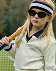 A girl with long blonde hair wears Okkia black sunglasses, a striped headband, and a cream ribbed tennis outfit. She holds a wooden racket over her shoulder on a tennis court with trees in the background.