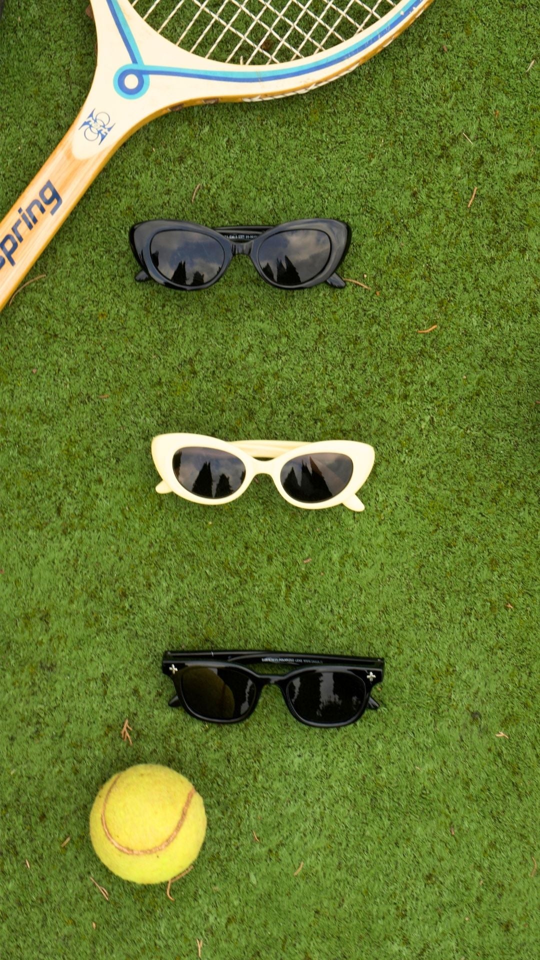 Three pairs of retro-style Okkia Kinderbrille in black, white, and black TPEE frames are arranged vertically on green grass with a tennis racket above and a yellow tennis ball below—a sporty, stylish composition.