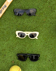 Three pairs of retro-style Okkia Kinderbrille in black, white, and black TPEE frames are arranged vertically on green grass with a tennis racket above and a yellow tennis ball below—a sporty, stylish composition.