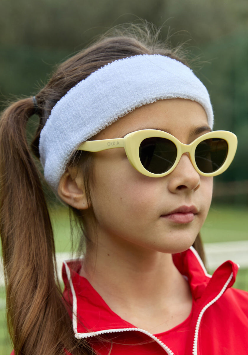 A young girl with long brown pigtails wears a white terry headband, cream Kinderbrille by Okkia, and a red zip-up jacket. Outdoors with a blurred green background, she gazes to the side with a calm expression.