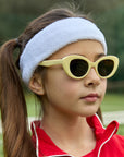 A young girl with long brown pigtails wears a white terry headband, cream Kinderbrille by Okkia, and a red zip-up jacket. Outdoors with a blurred green background, she gazes to the side with a calm expression.