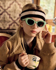 A young girl with long blond hair wears mint green Okkia sunglasses and a black-and-white headband. She holds a rooster teacup, raises a finger beside her glasses, and sits indoors in a tan jacket with patterned wallpaper behind her.