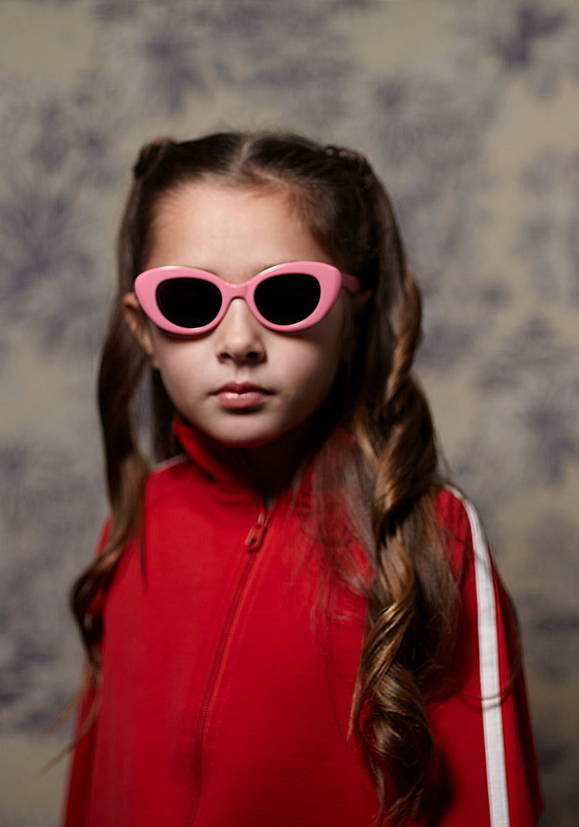 A young girl with long, wavy brown hair wears pink Okkia children’s sunglasses and a red track jacket with a white sleeve stripe, facing the camera seriously against a softly blurred Paris-inspired background.