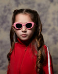 A young girl with long, wavy brown hair wears pink Okkia children’s sunglasses and a red track jacket with a white sleeve stripe, facing the camera seriously against a softly blurred Paris-inspired background.