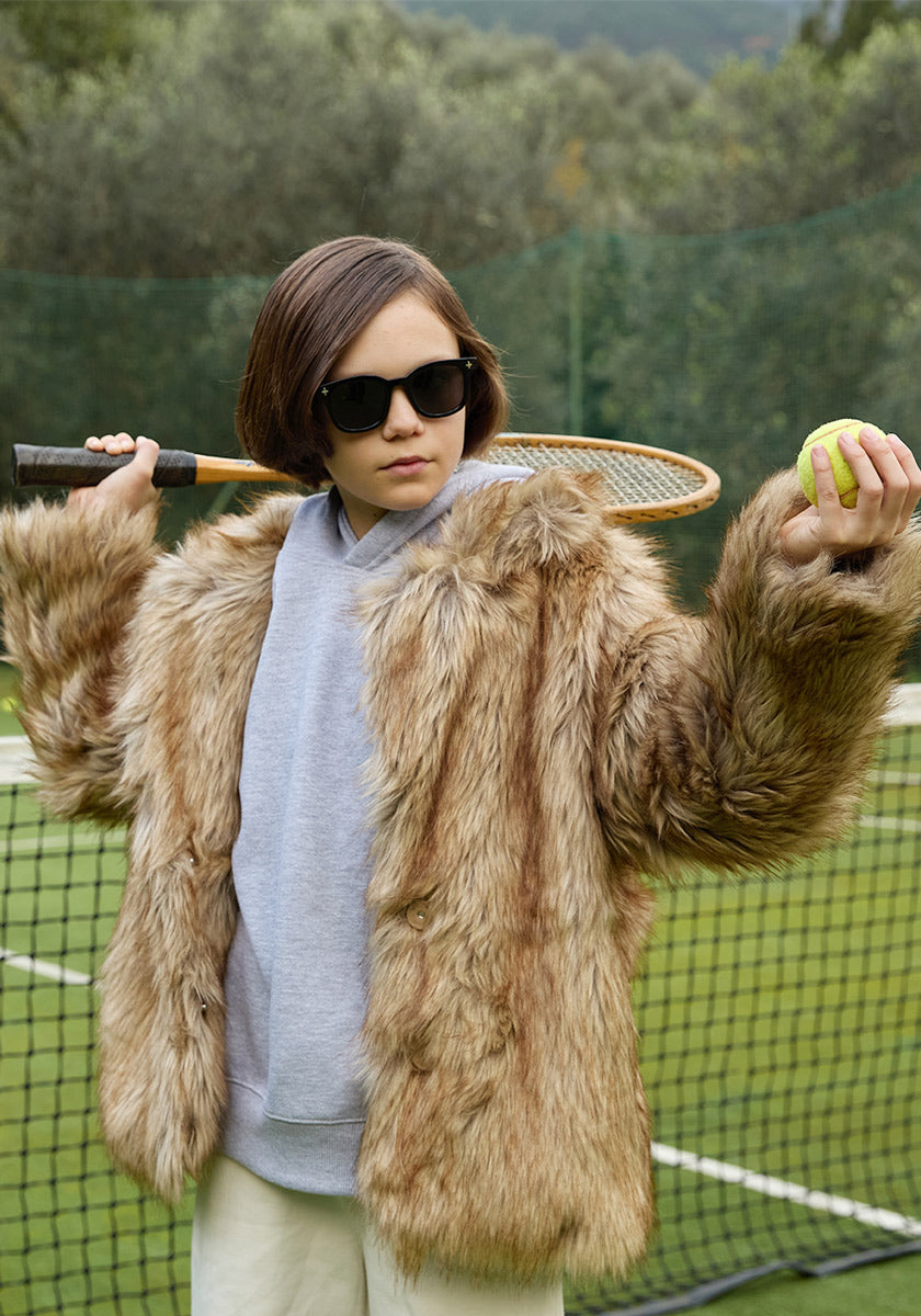 A young person with brown hair wears Okkia Kinder-Sonnenbrille and a furry brown coat over a gray hoodie, standing confidently on a tennis court. Tennis racket on one shoulder, ball in hand, framed by trees.