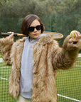 A young person with brown hair wears Okkia Kinder-Sonnenbrille and a furry brown coat over a gray hoodie, standing confidently on a tennis court. Tennis racket on one shoulder, ball in hand, framed by trees.