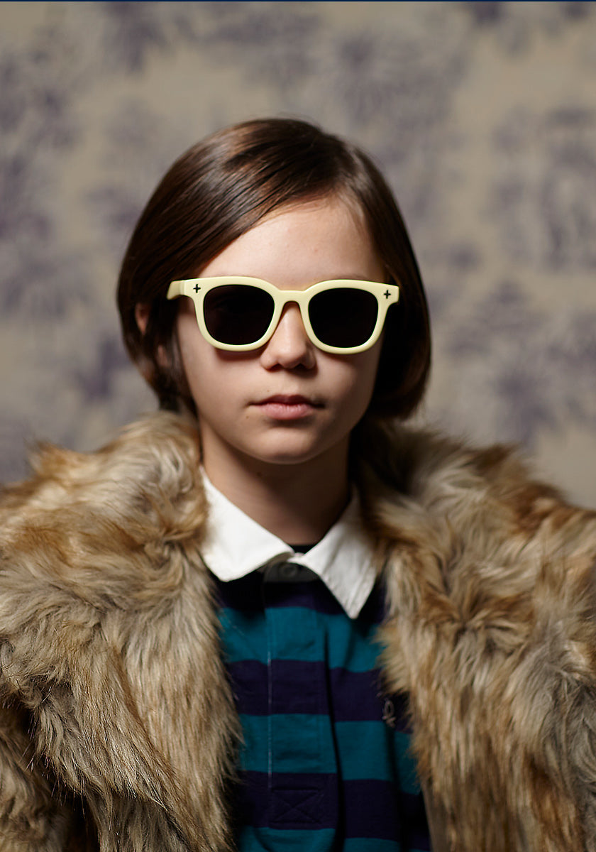 A young person with short brown hair wears cream-colored Okkia Kinder-Sonnenbrille aus flexiblem TPEE-Material, a fur coat, and a navy-green striped collared shirt, standing before a patterned backdrop with a neutral, direct expression.
