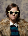A young person with short brown hair wears cream-colored Okkia Kinder-Sonnenbrille aus flexiblem TPEE-Material, a fur coat, and a navy-green striped collared shirt, standing before a patterned backdrop with a neutral, direct expression.