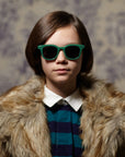A young person with straight brown hair wears Okkia grüne Kinder-Sonnenbrille, a fur coat, and a blue-and-navy striped shirt with a white collar, standing before floral wallpaper—perfekt für zuverlässigen Schutz.