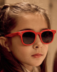 A young girl with long brown hair, teilweise zurückgebunden, trägt eine Okkia Kinder-Sonnenbrille aus flexiblem TPEE-Material und einen weißen Schal. Sie blickt ruhig zur Seite; der Hintergrund ist warm verschwommen.