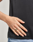 A person in a black shirt and light blue jeans rests their left hand on their hip, showing off neatly trimmed nails and wearing the Ring mit Herzen in Perlmutt-Optik by Princess Goes Hollywood among other rings.