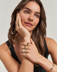 A woman with wavy brown hair wears a black sleeveless top and gold jewelry, including the Princess Goes Hollywood Vergoldeter Ring in Tropfenform. She rests her head on one hand, gazing softly upward with a relaxed expression.