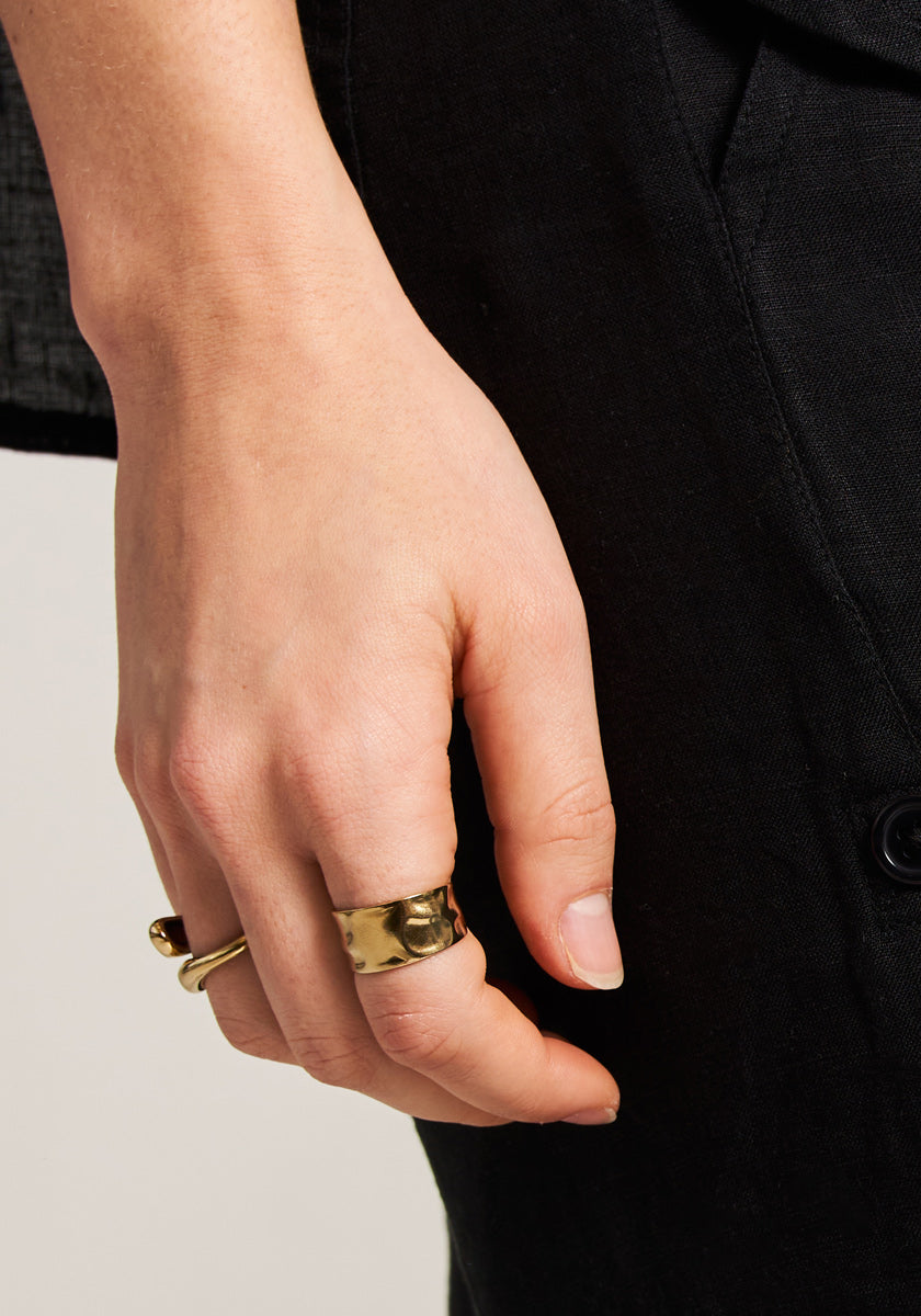 A close-up of a left hand by the side, wearing two chunky gold rings by Princess Goes Hollywood—one an open ring with 18K gold plating—on the middle finger. The black pants and dark jacket contrast against a neutral background.