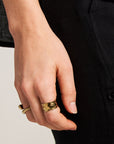 A close-up of a left hand by the side, wearing two chunky gold rings by Princess Goes Hollywood—one an open ring with 18K gold plating—on the middle finger. The black pants and dark jacket contrast against a neutral background.