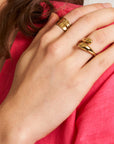 A close-up shows a hand wearing the Princess Goes Hollywood Vergoldeter Ring in Tropfenform, crafted from Edelstahl 304 with 18K Vergoldung, gently resting on a shoulder. The person wears bright pink, brown hair partly visible against a neutral background.