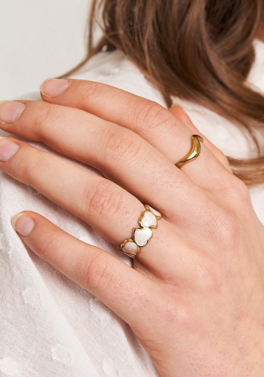A left hand with neatly trimmed nails wears two rings: a gold open ring on the index finger and Princess Goes Hollywoods Ring mit Herzen in Perlmutt-Optik on the ring finger. The hand rests on white textured fabric.
