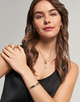 A woman with wavy brown hair wears a black sleeveless top and gold jewelry from Princess Goes Hollywood, including a Retro-Stil Halskette with rechteckiger Anhänger, hoop earrings, layered necklaces, a ring, and bracelet. She smiles confidently at the camera.