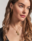 A woman with wavy light brown hair wears gold hoop earrings and two layered Princess Goes Hollywood necklaces with rectangular stainless steel pendants. She has green eyes, natural makeup, a slight smile, a black top, and looks off to the side.