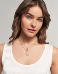 A woman with wavy brown hair, wearing a white sleeveless top and a Princess Goes Hollywood Retro-Stil Halskette mit rechteckigem Anhänger, poses softly. She has light skin, subtle makeup, and gold hoop earrings against a light gray background.