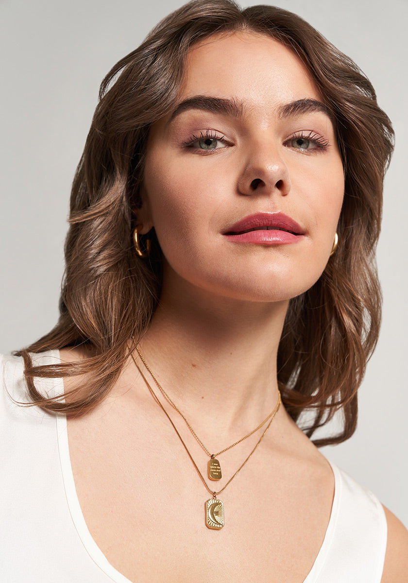 A woman with wavy brown hair and light skin wears gold hoop earrings and a Princess Goes Hollywood Retro-Stil Halskette with two layered pendants. In a white tank top, she smiles softly and gazes upward against a neutral background.