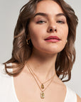 A woman with wavy brown hair and light skin wears gold hoop earrings and a Princess Goes Hollywood Retro-Stil Halskette with two layered pendants. In a white tank top, she smiles softly and gazes upward against a neutral background.