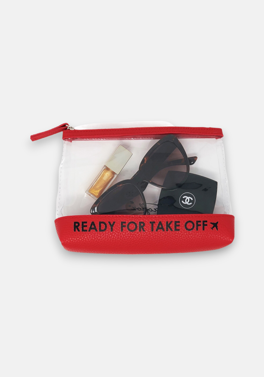 A clear Frogbox by Princess Kulturbeutel with red trim and bold “READY FOR TAKE OFF” text holds black Chanel sunglasses in their case and a gold-capped lipstick, set against a plain white background.