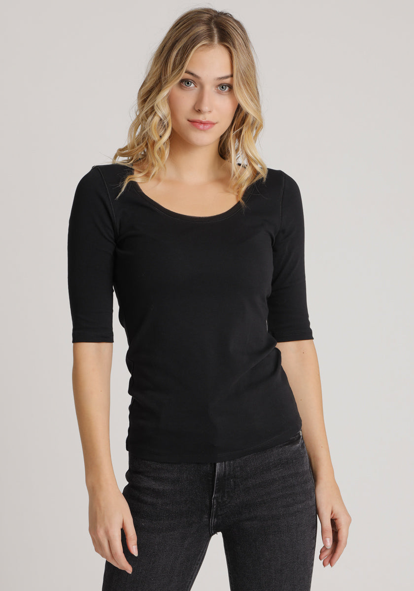 A young woman with wavy blonde hair wears a fitted black Princess Goes Hollywood Baumwolle shirt with 3/4-Arm sleeves and black jeans, standing against a plain light gray background and looking at the camera neutrally.