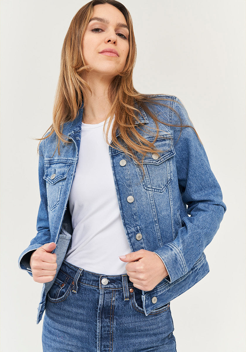 Eine Person mit langem, hellbraunem Haar trägt eine offene blaue Jeansjacke von Princess Goes Hollywood aus Baumwolle mit Brusttaschen und Hemdkragen über weißem T-Shirt und Jeans, steht selbstbewusst vor hellem Hintergrund und blickt leicht nach oben.