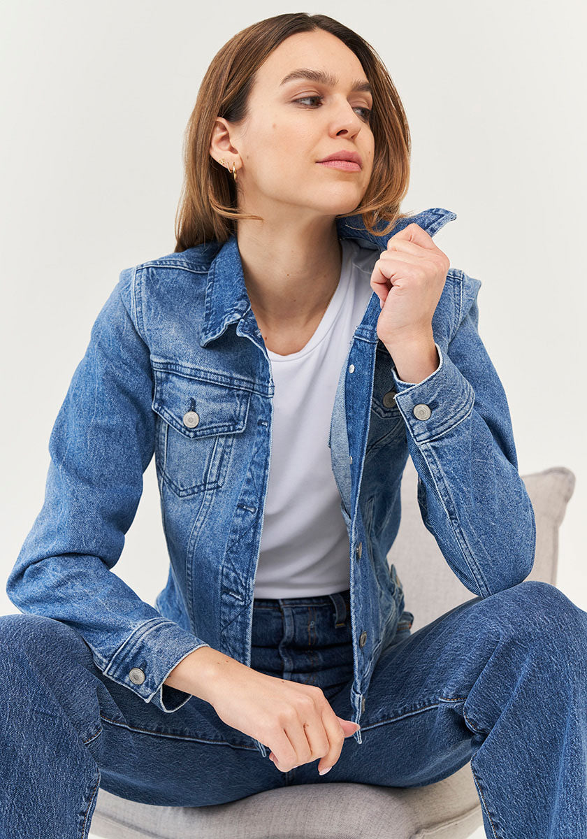 A woman with straight brown hair models a blue denim jacket by Princess Goes Hollywood, featuring Brusttaschen and Hemdkragen, layered over a white T-shirt and matching jeans. She sits on a light chair, confidently holding her collar against a neutral backdrop.