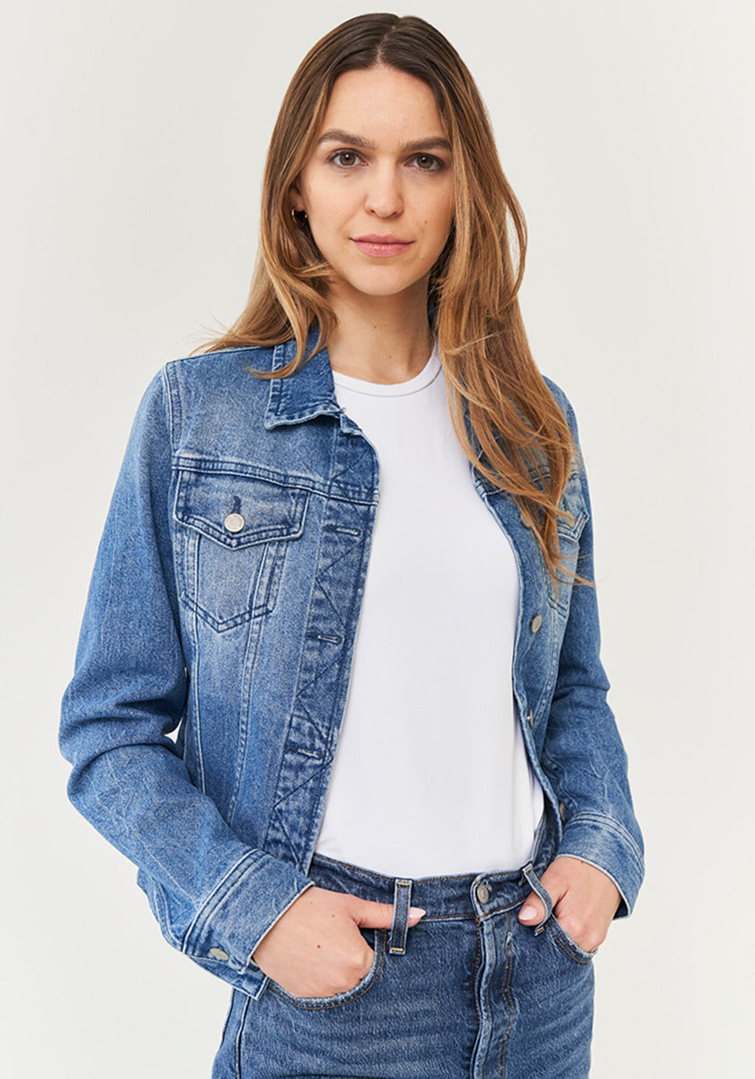 A woman with long, straight brown hair wears a Princess Goes Hollywood blue denim jacket with Hemdkragen and Brusttaschen over a white t-shirt and blue jeans, standing confidently with hands in her jacket pockets against a plain light background.