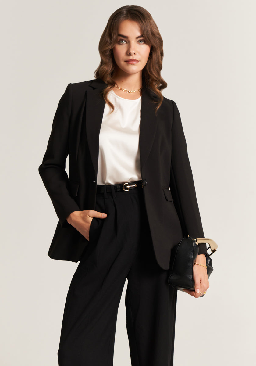 A woman with wavy brown hair wears a Princess Goes Hollywood schwarzer Blazer aus stretchigem Webstoff und eine schmal geschnittene Hose mit weißer Bluse. Sie hält eine schwarze Handtasche, blickt selbstbewusst in die Kamera.