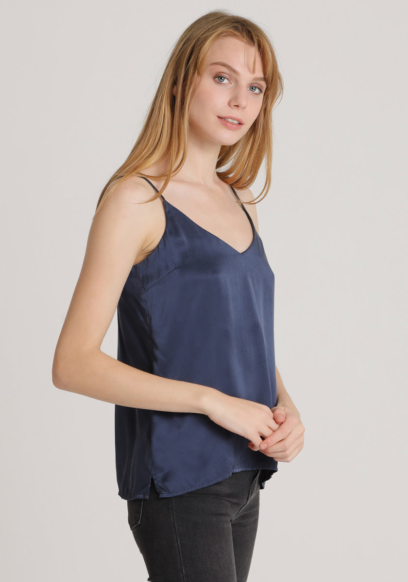 A young woman with long blonde hair wears a Princess Goes Hollywood dark blue satin camisole top with V-neck and thin straps, paired with black jeans. She stands against a light background, facing slightly sideways and smiling gently at the camera.