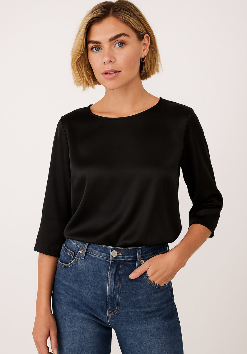 Eine Frau mit kurzem blondem Bob trägt eine schwarze Seidenbluse mit 3/4-Ärmeln und lockerem Schnitt von Princess Goes Hollywood, in hoch taillierte blaue Jeans gesteckt. Sie steht vor hellem Hintergrund, blickt neutral und hat eine Hand in der Tasche.