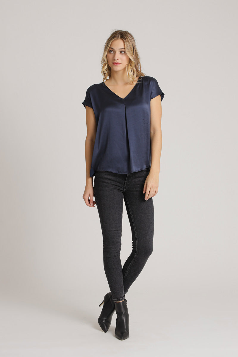 A woman with wavy blonde hair models a Princess Goes Hollywood navy V-neck blouse, black skinny jeans, and black ankle boots against a light gray background. She stands relaxed, one leg bent and arms at her sides.