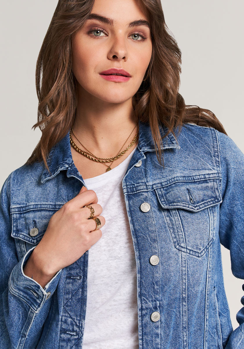 A confident woman with wavy brown hair and green eyes wears a light blue Princess Goes Hollywood Denimjacke with Hemdkragen and Brusttaschen over a white shirt, accessorized with gold jewelry, posing against a neutral background.