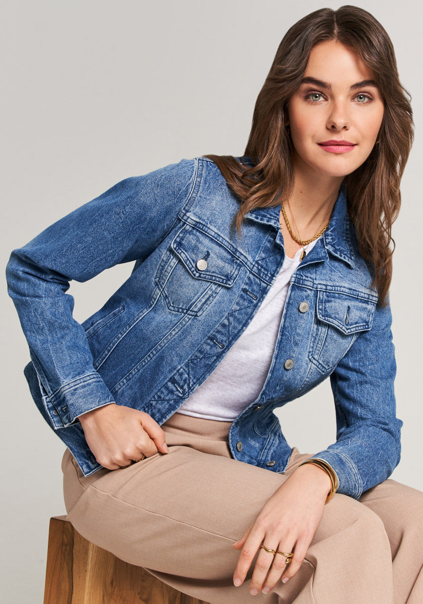 A woman with wavy brown hair sits on a wooden stool, wearing a blue denim jacket with Hemdkragen and Brusttaschen by Princess Goes Hollywood over a white shirt and beige pants. She smiles, her outfit crafted from comfortable Baumwolle.