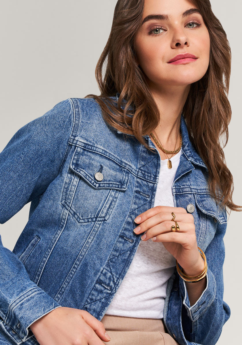 A woman with wavy brown hair models a blue denim jacket by Princess Goes Hollywood with Hemdkragen and Brusttaschen over a white top. She wears gold jewelry, smiles subtly, and poses hand near her chin against a neutral background.