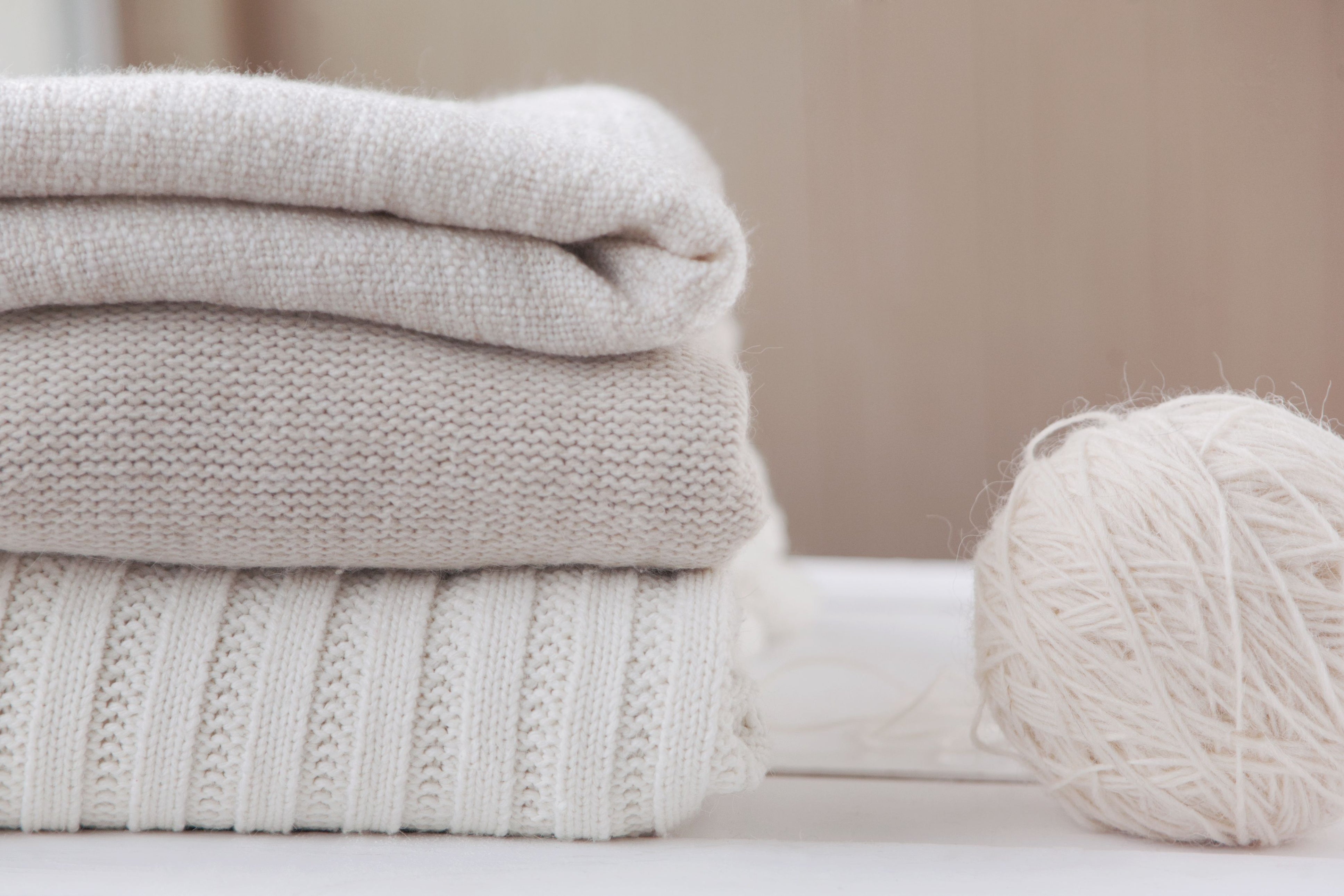 Three folded sweaters in shades of cream and beige are stacked on a white surface. To the right, a ball of light cream-colored yarn sits, adding a cozy, soft texture to the scene. The background is softly blurred, emphasizing the knits and yarn.