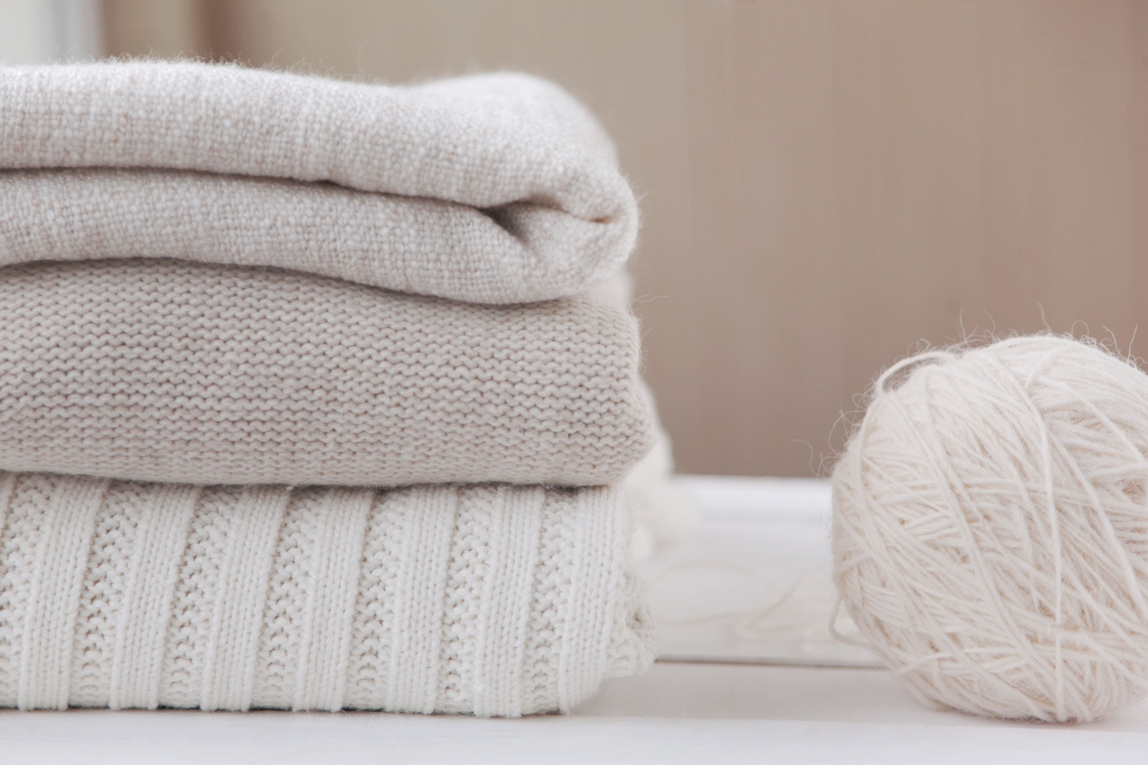 Three folded sweaters in shades of cream and beige are stacked on a white surface. To the right, a ball of light cream-colored yarn sits, adding a cozy, soft texture to the scene. The background is softly blurred, emphasizing the knits and yarn.
