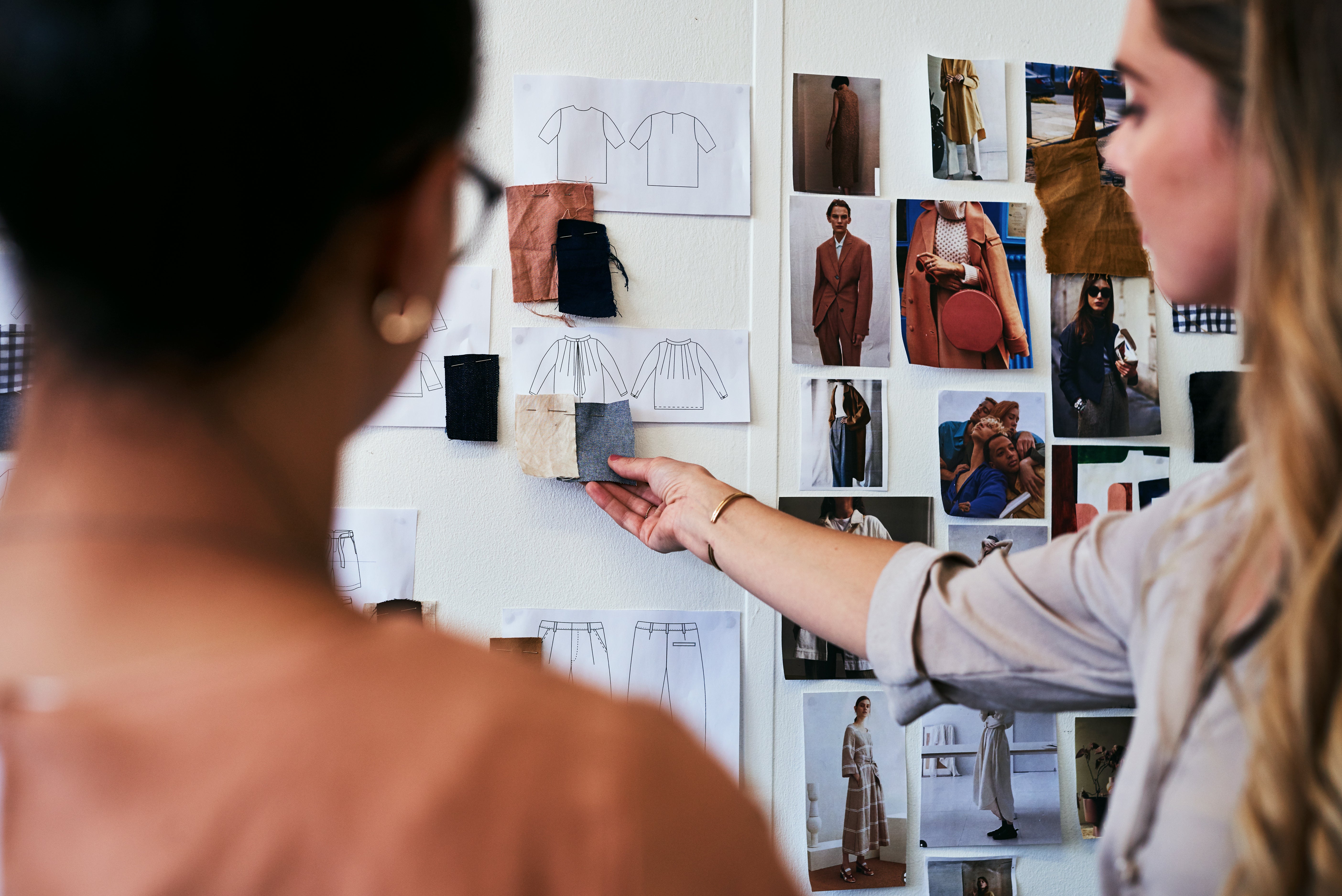 Two women stand facing a wall covered in fashion sketches, fabric swatches, and photos of models in various outfits. One woman points to fabric samples pinned near clothing sketches, suggesting a discussion about fashion design or garment planning.