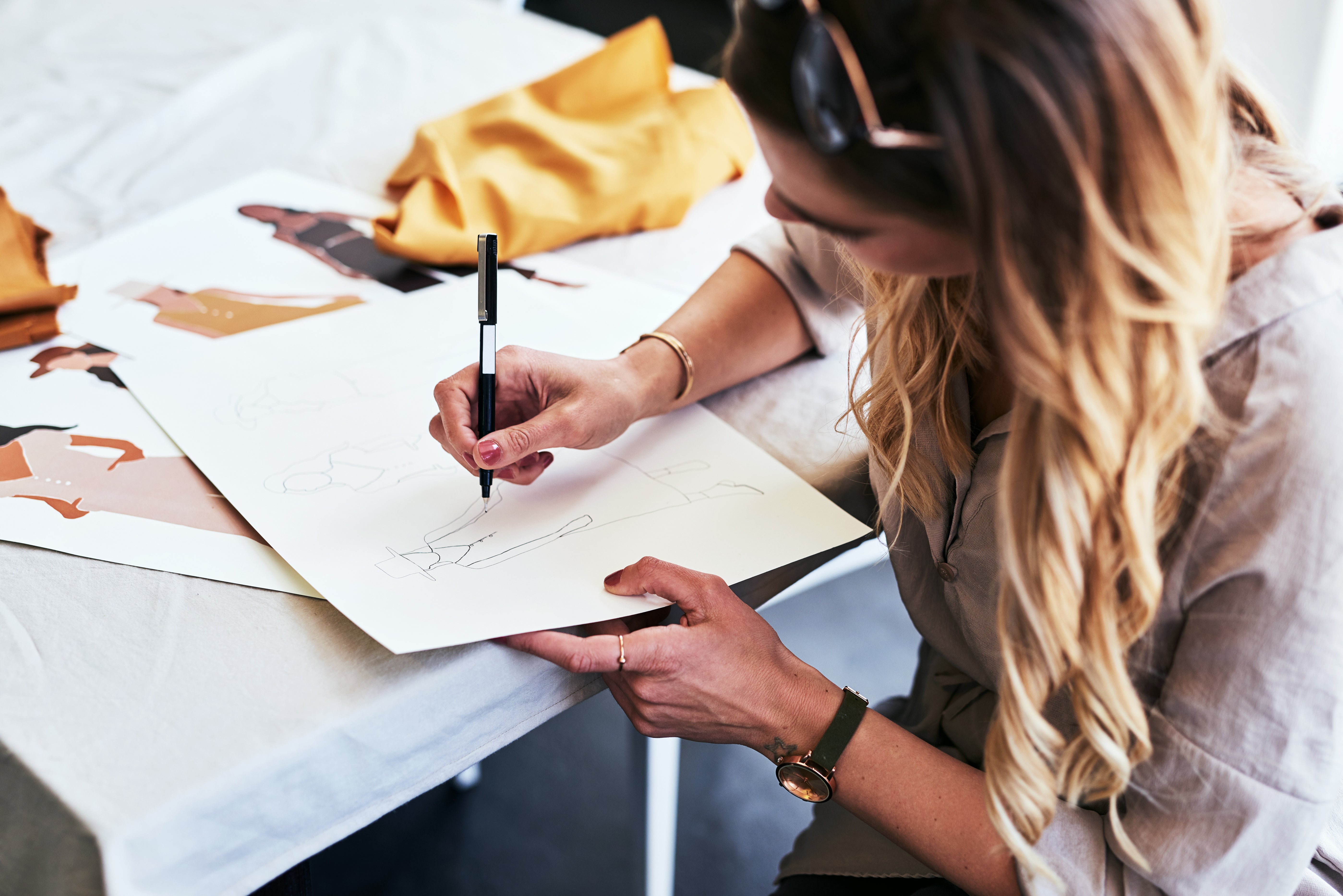 A woman with long blonde hair sketches fashion designs on white paper at a table. She holds a pen and wears a watch and beige blouse. Nearby are more fashion sketches and a piece of mustard-yellow fabric. The setting appears creative and focused.