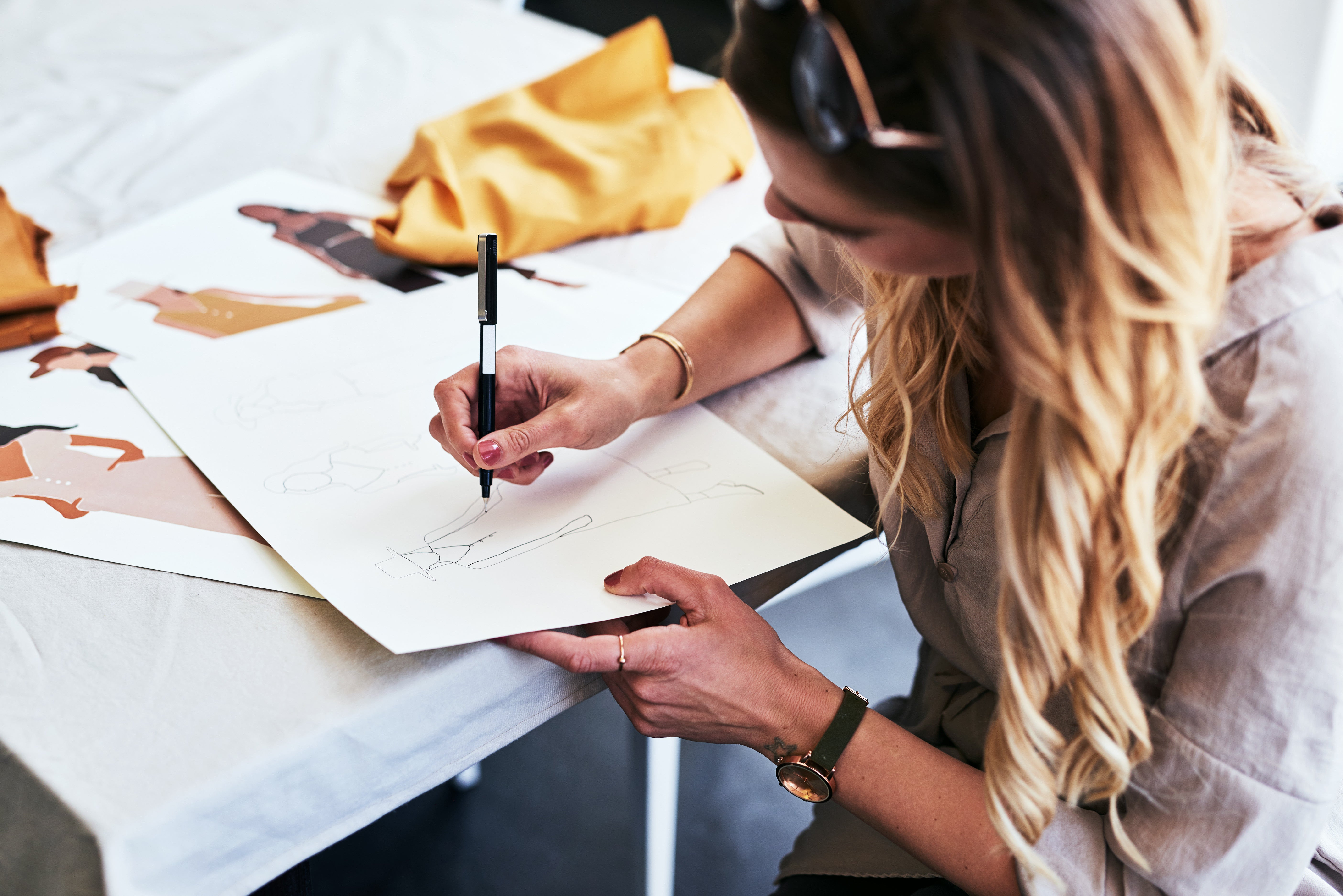 A woman with long blonde hair sketches fashion designs on white paper at a table. She holds a pen and wears a watch and beige blouse. Nearby are more fashion sketches and a piece of mustard-yellow fabric. The setting appears creative and focused.