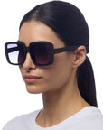A woman with long dark hair tied back wears Okkia oversized sunglasses with gradient lenses and brand name on the arm. She has a neutral expression, dressed in a white shirt against a plain white background.