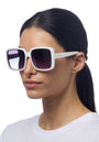 A woman with straight dark hair tied back, wearing a white top and Okkia oversized square sunglasses with dark gradient lenses and UV 400 protection, stands against a plain white background, facing slightly to the right with a neutral expression.