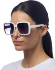 A woman with straight dark hair tied back, wearing a white top and Okkia oversized square sunglasses with dark gradient lenses and UV 400 protection, stands against a plain white background, facing slightly to the right with a neutral expression.