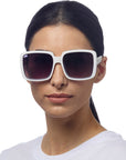 A woman with straight dark hair tied back wears large Okkia Sonnenbrille with dark UV 400 lenses. She has a neutral expression, is dressed in a plain white shirt, and the background is plain white.
